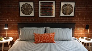 Ein Schlafzimmer mit Backsteinwand, einem Bett mit gestreifter Bettwäsche, zwei Nachttischen mit Lampen und drei gerahmten Wandbildern.