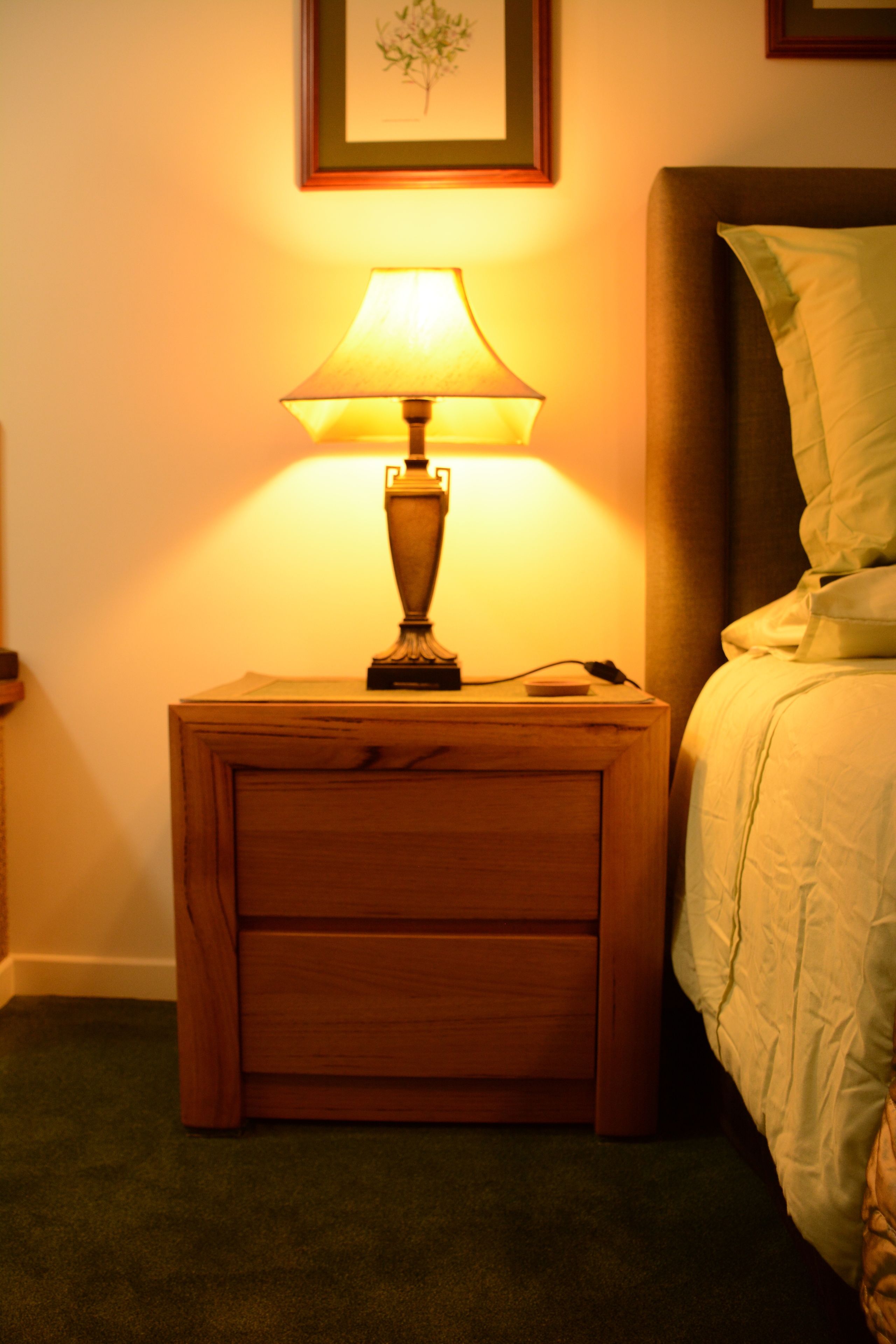 A wooden bedside table with a lamp, a framed picture on the wall, and a bed with a green pillow.