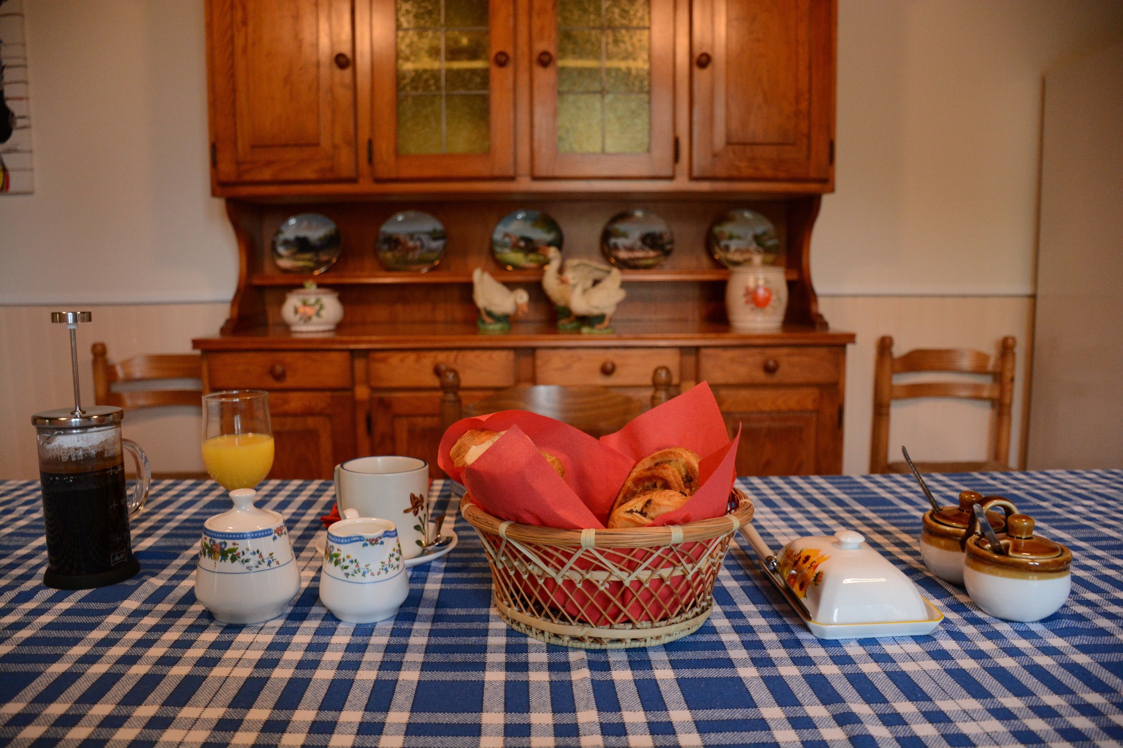 A checkered tablecloth, a coffee pot, a glass of orange juice, a basket of bread, and ceramic containers on a table.
