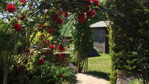 Un sentier de jardin agrémenté d’une tonnelle couverte de roses, de murs en pierre et de diverses plantes.