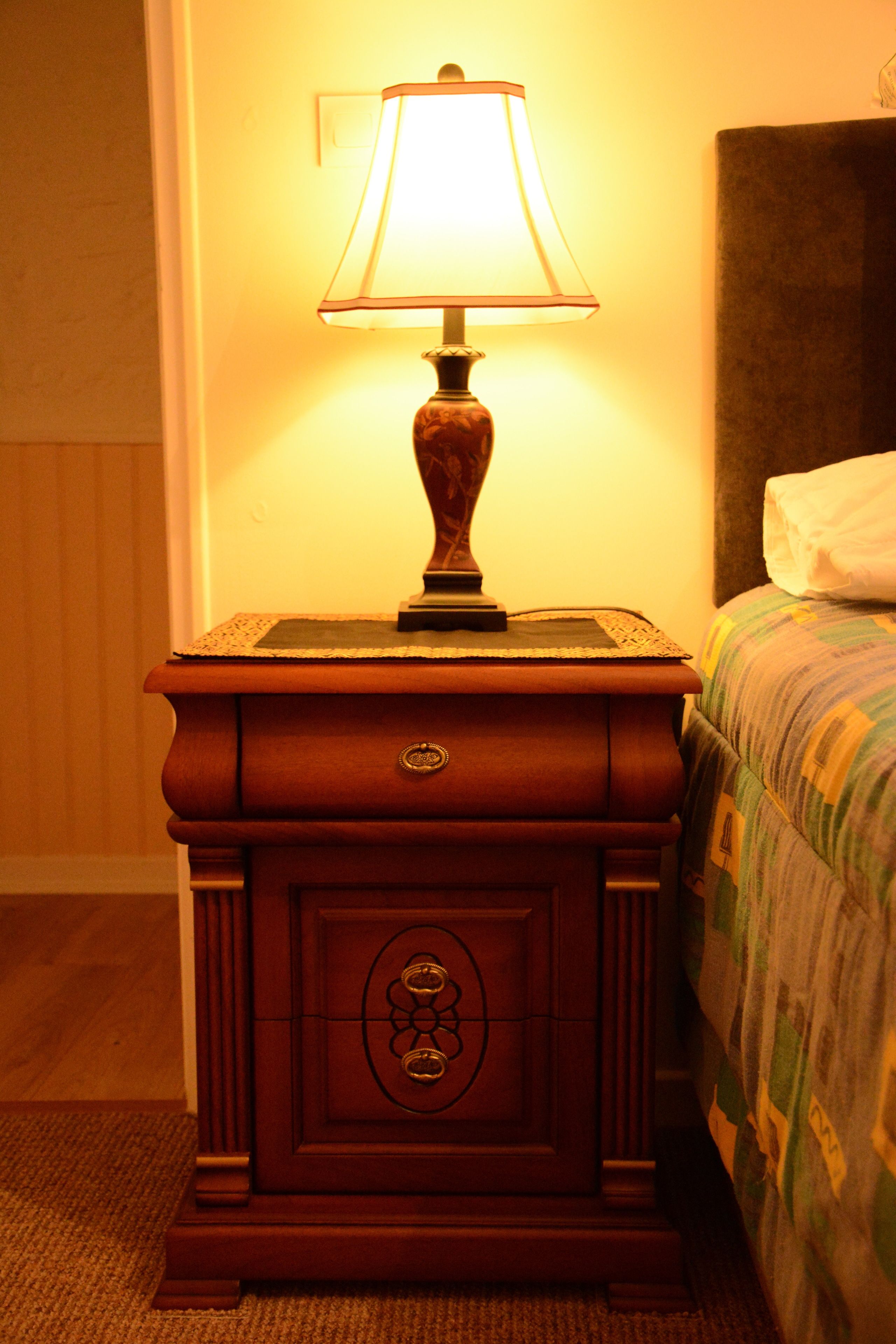 Uma mesa de cabeceira de madeira com uma luminária, uma cama com colcha estampada e uma cabeceira.