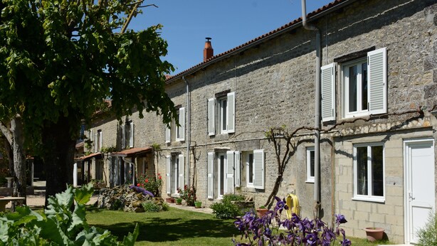 Un bâtiment en pierre avec des volets blancs, un jardin fleuri et un ciel dégagé.