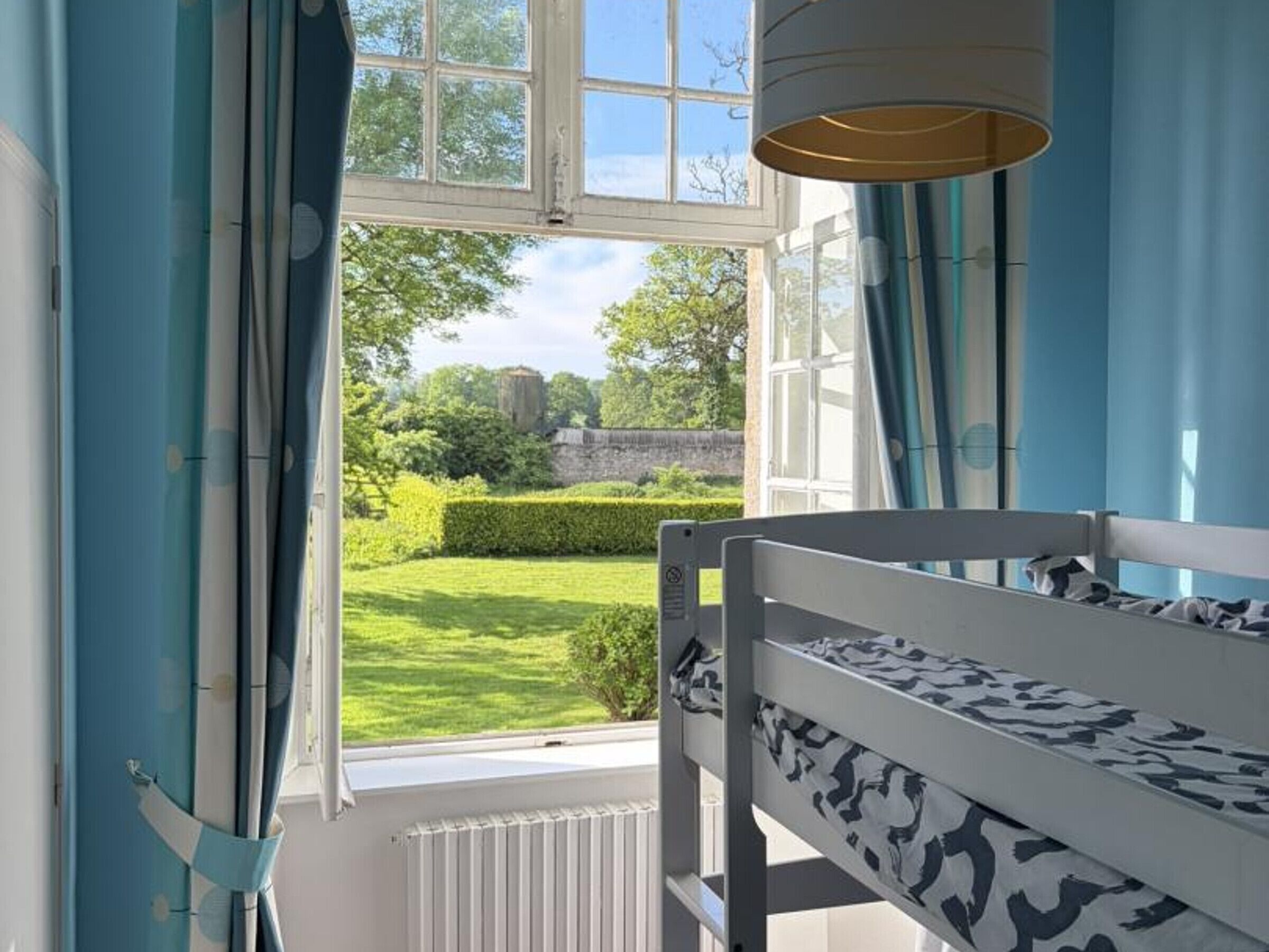 A bunk bed with a patterned mattress, a window with curtains, and a view of a garden and stone wall.
