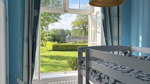 Ein Stockbett mit gemustertem Bettzeug, ein Fenster mit Vorhängen und Blick auf einen Garten und eine Steinmauer.