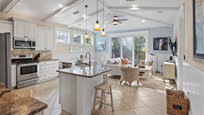 A spacious kitchen with white cabinetry, a central island, and a dining area with wicker furniture. - Minnie Cottage- Beautiful 3 bedroom heated pool/spa and short walk to the beach (Holmes Beach)