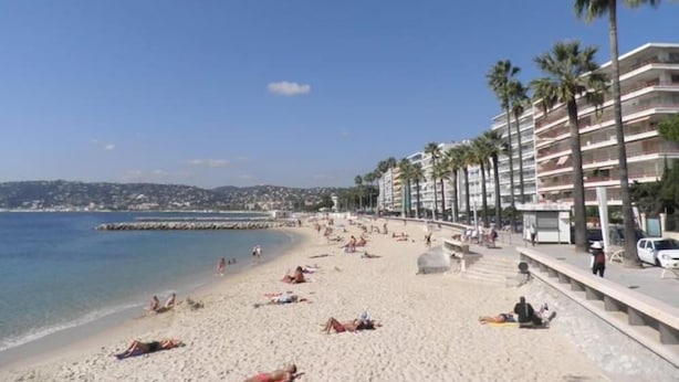 A beach with people sunbathing and swimming, a clear sky, and a row of tall buildings with palm trees.