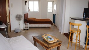 A bedroom with a single bed, a small table with a basket, a chair, a fan, and a desk with a chair.