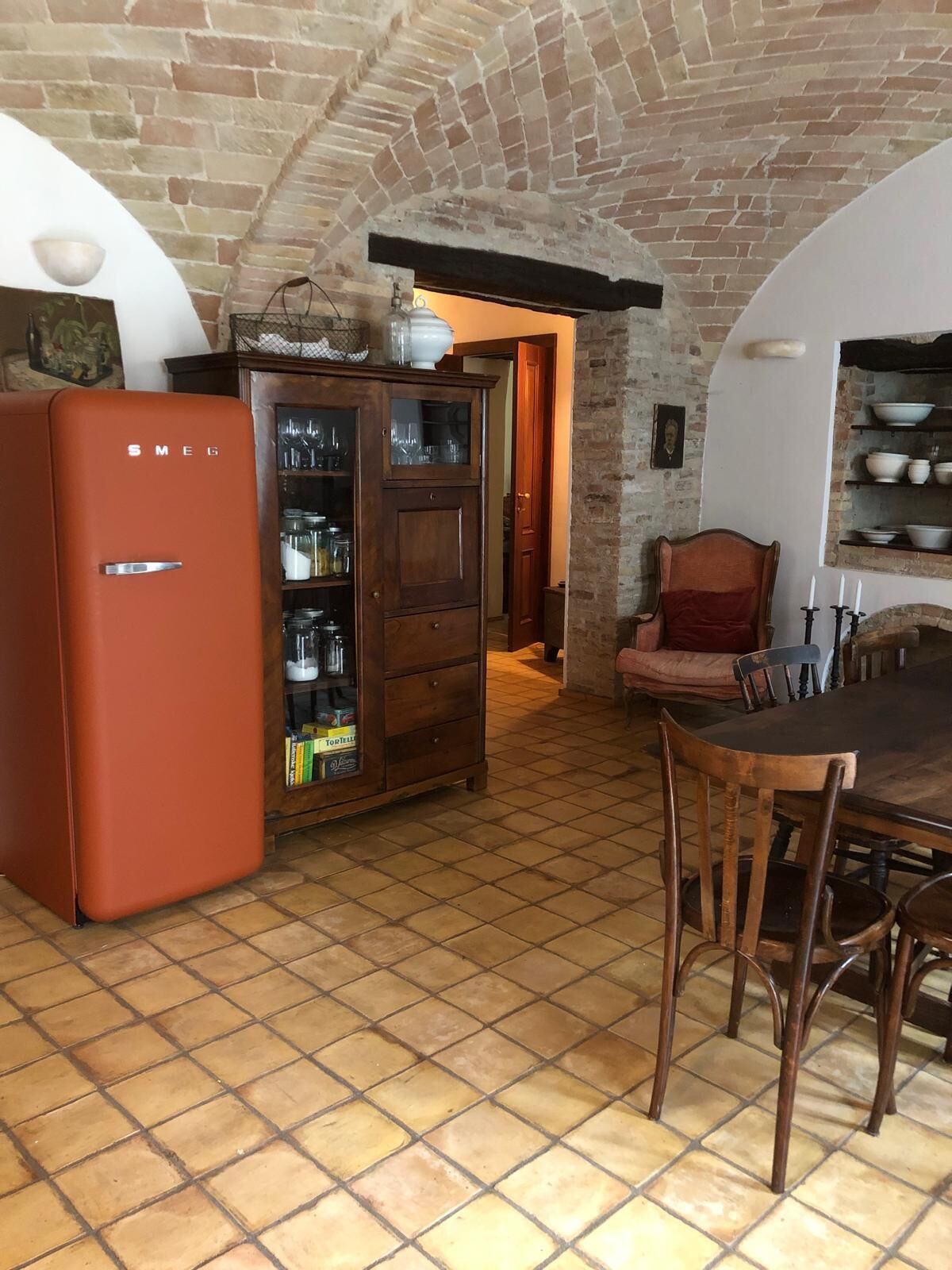 A kitchen with a tiled floor, a wooden cabinet with glass doors, a vintage orange refrigerator, and a dining table with chairs.