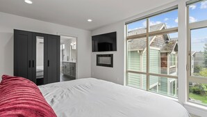 A bedroom with a large bed, a TV mounted on the wall, and a window with a view of neighboring houses. - New centrally located townhouse with rooftop deck. Walk to restaurants and shops (Seattle)