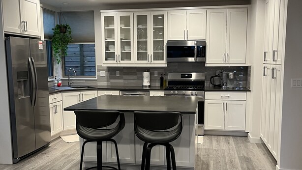 A modern kitchen with white cabinetry, a stainless steel refrigerator, a microwave, and a gas stove. There are two black bar stools at the island. - New centrally located townhouse with rooftop deck. Walk to restaurants and shops (Seattle)