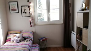 A bedroom with a bed, a window with curtains, a bedside table with books, and a shelf with decorative items. - Bohemian chic apartment in the heart of the Antigone district (Montpellier)
