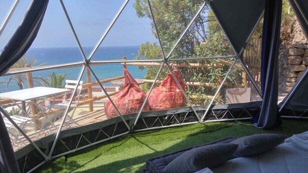A geodesic dome room with a view of the sea, a small outdoor seating area, and a bed with pillows. - Eremo di Montevergine : Seaview Dome, suspended between sky and sea (Forio)