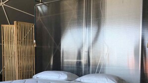 A bed with white bedding and a wooden platform, a bamboo screen, and a metal mesh partition. - Eremo di Montevergine : Seaview Dome, suspended between sky and sea (Forio)