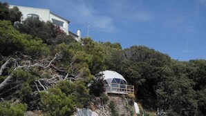 A dome-shaped structure with a wooden deck, surrounded by dense forest and a white building in the background. - Eremo di Montevergine : Seaview Dome, suspended between sky and sea (Forio)