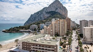 Eine Küstenstadtlandschaft mit einem markanten Berg im Hintergrund, modernen Hochhäusern, einem Strand und wolkenlosem Himmel.