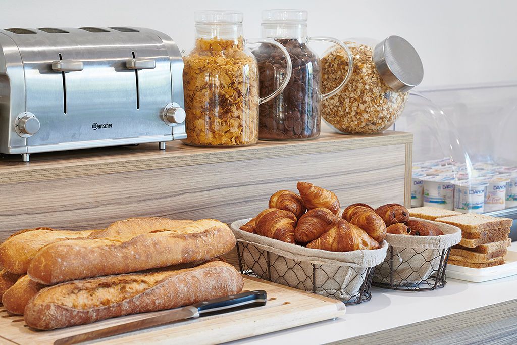 Un plan de travail de cuisine avec un grille-pain, des bocaux de nourriture, un panier de croissants et une planche à découper avec un couteau.
