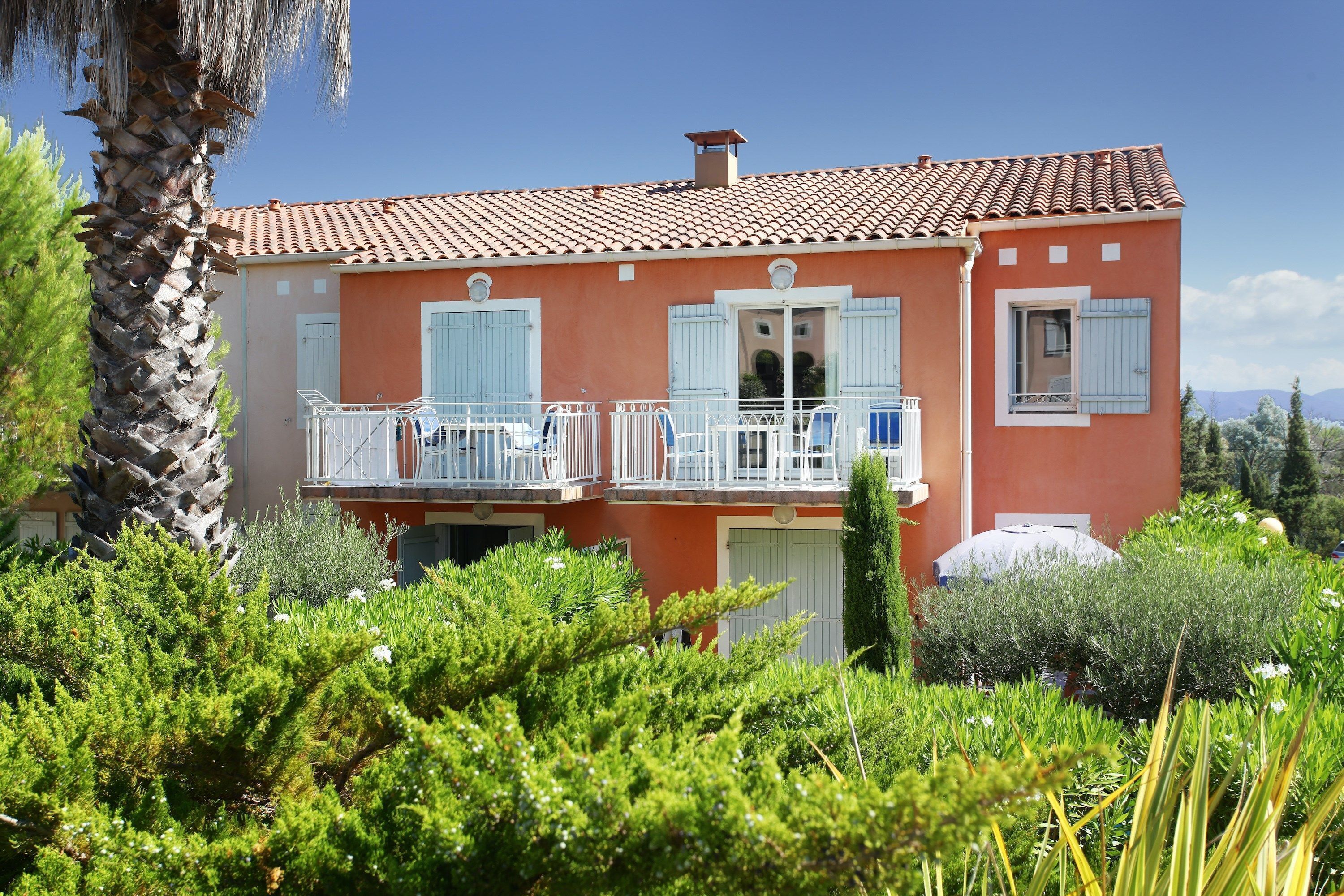 Une maison à deux étages, avec un toit recouvert de tuiles rouges, des volets blancs et un balcon doté de balustrades blanches.