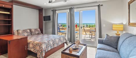 A bedroom with a bed, a desk, a sofa, a ceiling fan, and a view of the beach through the sliding doors.