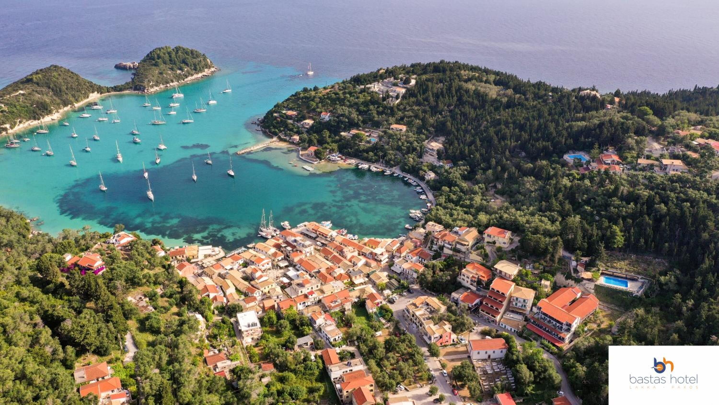 An aerial view of a coastal town with a marina, houses with red roofs, and a clear blue sea.