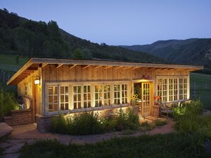 Une cabane en bois dotée de grandes fenêtres, d’une fondation en pierre et d’un porche avec un banc.