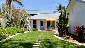 Un jardin bien entretenu avec une pelouse, des pas japonais, une terrasse avec des chaises et un parasol jaune.