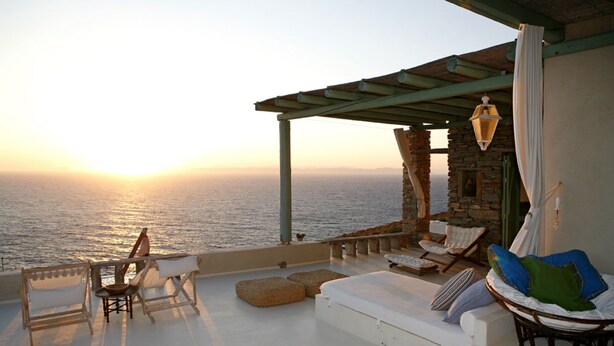 Une terrasse offrant une vue sur la mer au coucher du soleil, avec un canapé, des fauteuils et un fauteuil suspendu.