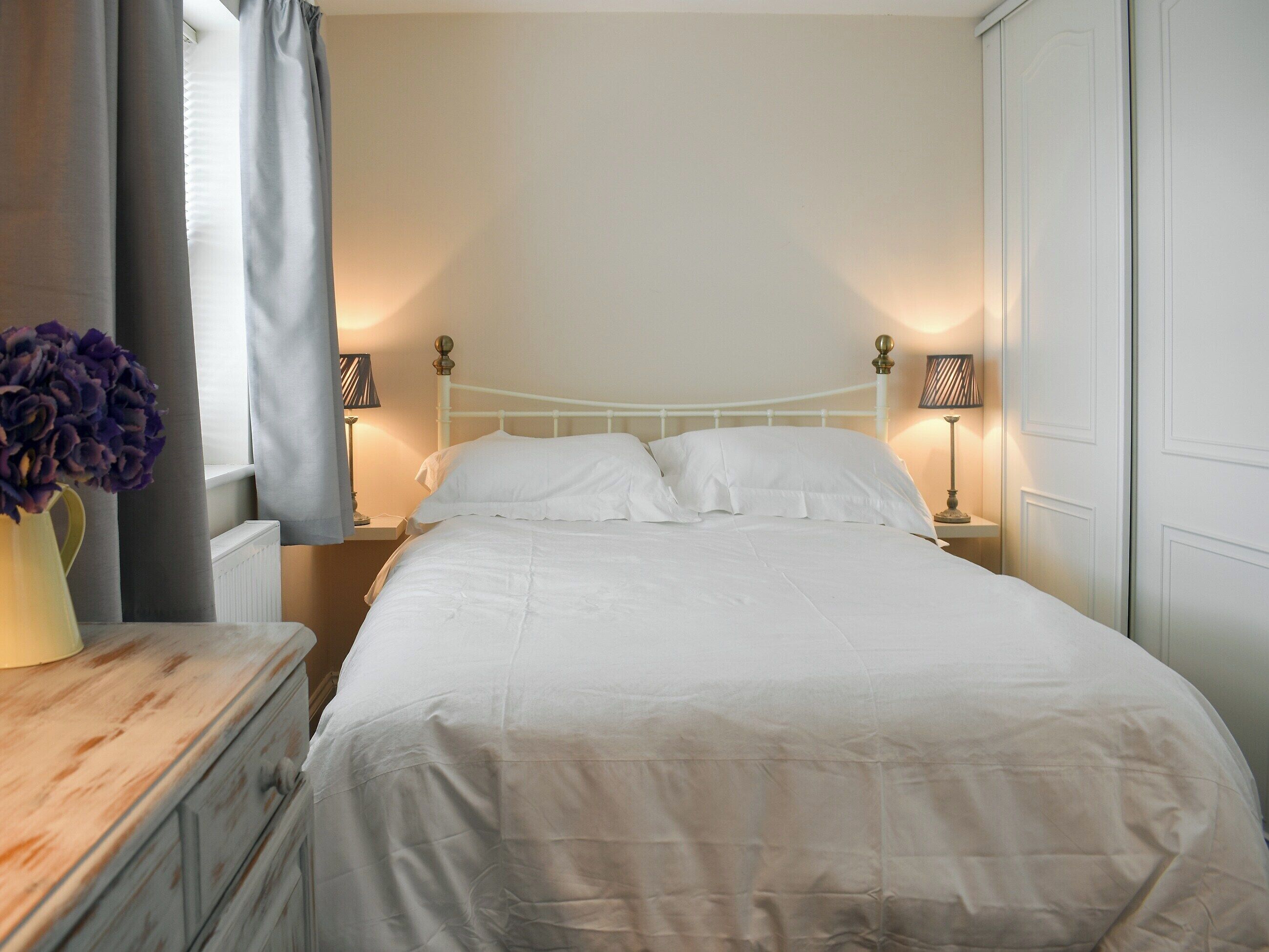 A neatly made bed with white linens, two bedside lamps, and a vase of purple flowers on a wooden nightstand.