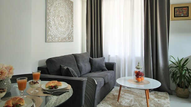 A modern living room with a grey sofa, a small round table with a glass jar of fruit, and a dining area with plates of food and drinks. - Split Centre VIVERE Apartment (Split)