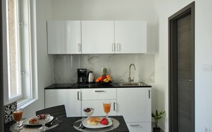 A compact kitchen with white cabinets, a black countertop, a coffee maker, a fruit bowl, and a dining table with breakfast items. - Split Centre VIVERE Apartment (Split)