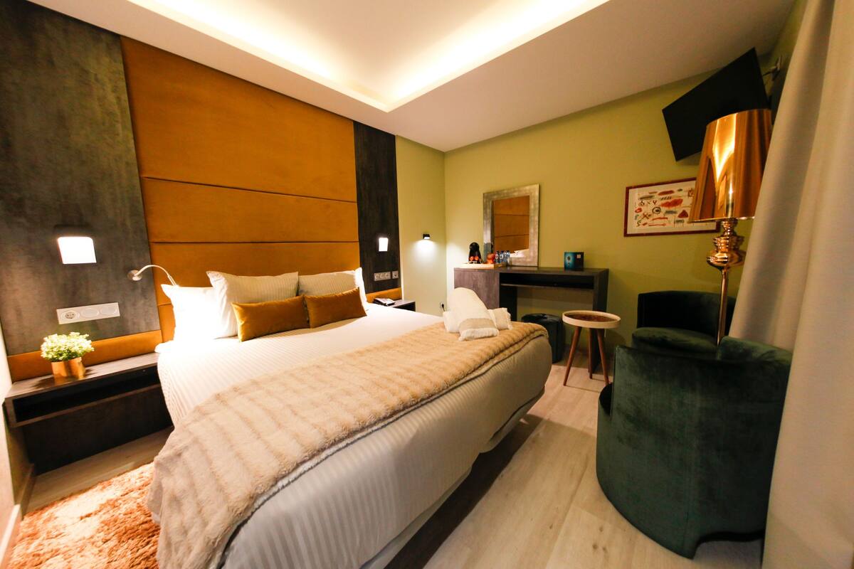 Habitación de hotel moderna con una cama grande, un sillón verde, una mesita y un televisor montado en la pared.