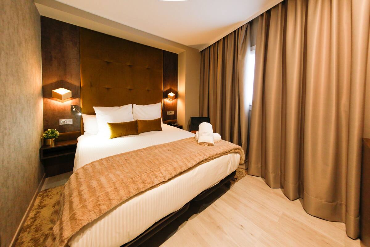 Una habitación de hotel con una cama grande, cabecera con iluminación integrada, mesita de noche con lámpara y ventana con cortinas.