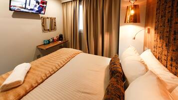 A hotel room with a bed, a TV mounted on the wall, a desk with a lamp, and curtains on the window.