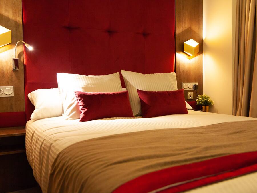 Quarto de hotel com uma cama grande, cabeceira vermelha e duas luminárias de leitura fixadas na parede.