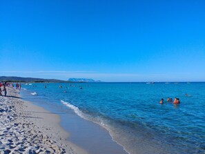 Una playa con agua azul cristalina, gente nadando y caminando por la orilla, y barcos a lo lejos.