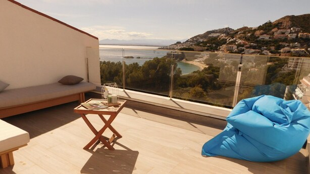 A balcony with a wooden floor, a glass table, a blue bean bag, and a sofa. - Modern house overlooking the sea, near the beach in Almadraba, Roses (ROSES)