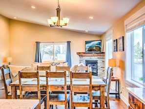 A dining area with a wooden table and chairs, a chandelier, a fireplace, and large windows.