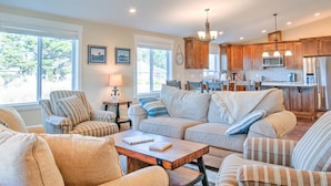 A spacious living room with a large sofa, armchairs, and a coffee table. There is a kitchen area with wooden cabinets and modern appliances in the background.