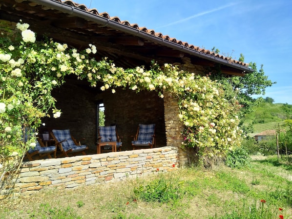 Un edificio de piedra con un porche cubierto, sillas de madera y enredaderas con flores blancas.