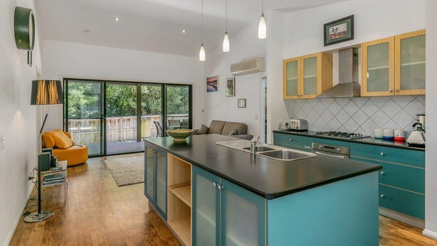Cocina moderna con gabinetes color esmeralda, encimera negra y una isla grande con un lavamanos. Hay un comedor con sofá y mesa, y un ventanal con vistas al exterior.