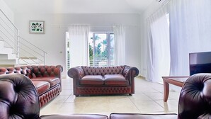 Un salón con un sofá de cuero marrón, una mesa de centro de madera y un televisor. Se ve una escalera a la izquierda, y una ventana con cortinas a la derecha.