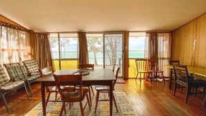 A dining area with a table and chairs, a sofa, and a view of the beach through large windows. - Stafford Hall - Burrum Heads- Riverfront- 3BR- Great Views (BURRUM HEADS)