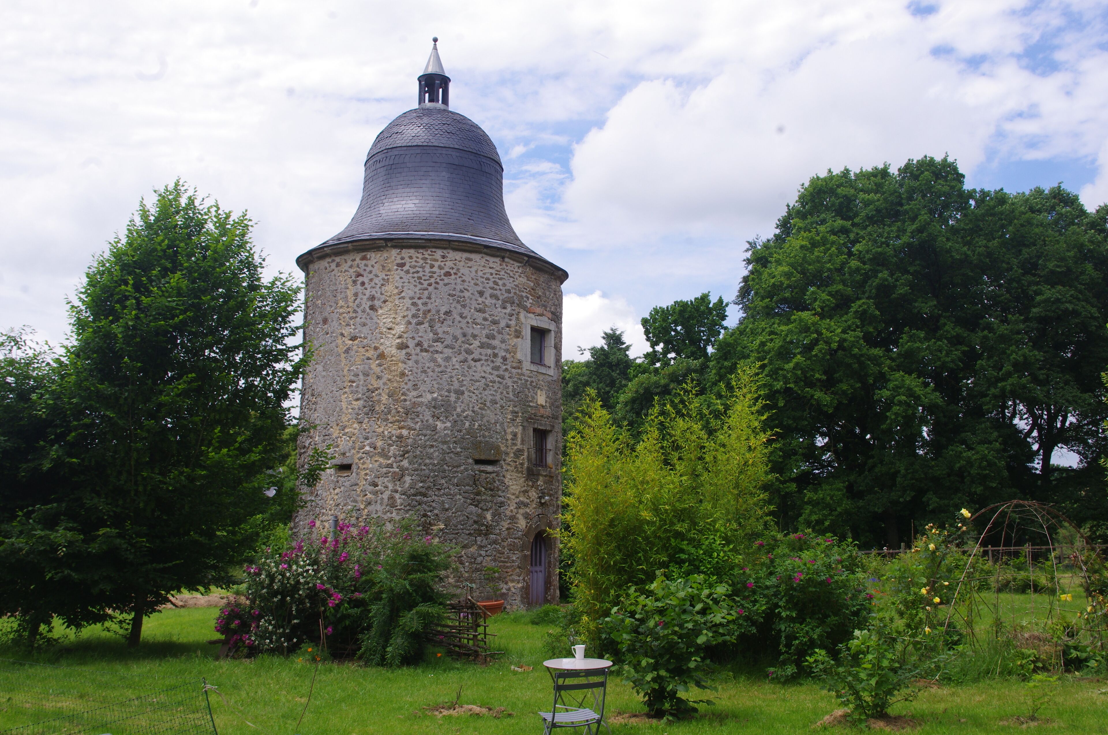 A stone tower with a conical roof, surrounded by lush greenery and a small garden with a table and chair.