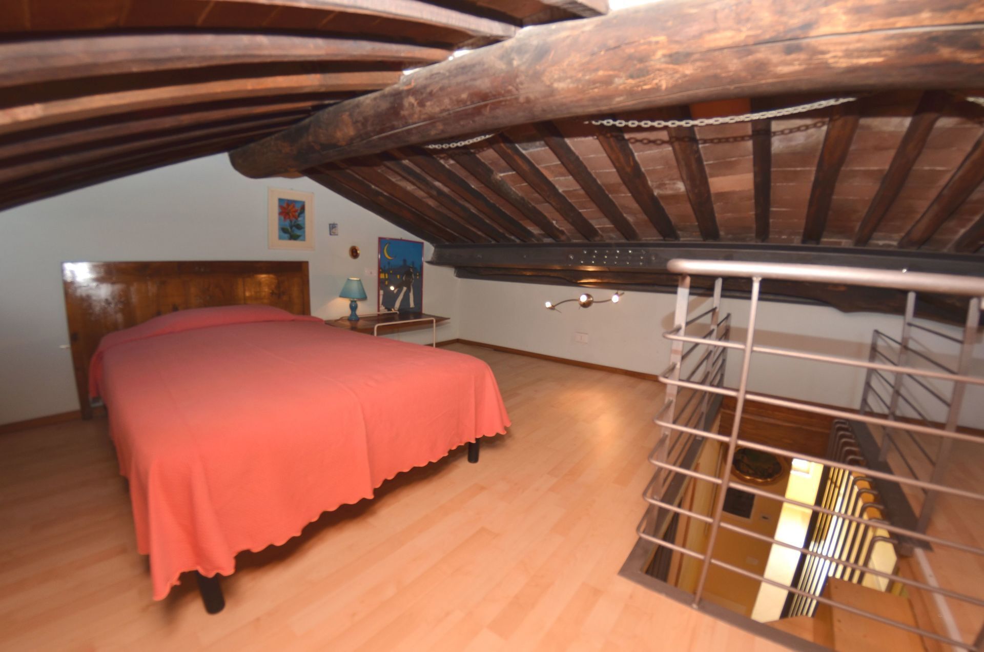 A bedroom with a wooden ceiling, a bed with a pink bedspread, a nightstand, and a wall-mounted shelf.