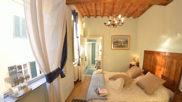 A bedroom with a bed, a nightstand, a chandelier, a window with curtains, and a door leading to a bathroom. - Beautiful apartment for 7 people with A/C, WIFI, TV and panoramic view (Lucca)
