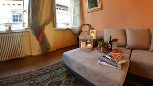 A cozy living room with a sofa, a table with books and a bottle, a window with curtains, and a radiator. - Beautiful apartment for 7 people with A/C, WIFI, TV and panoramic view (Lucca)