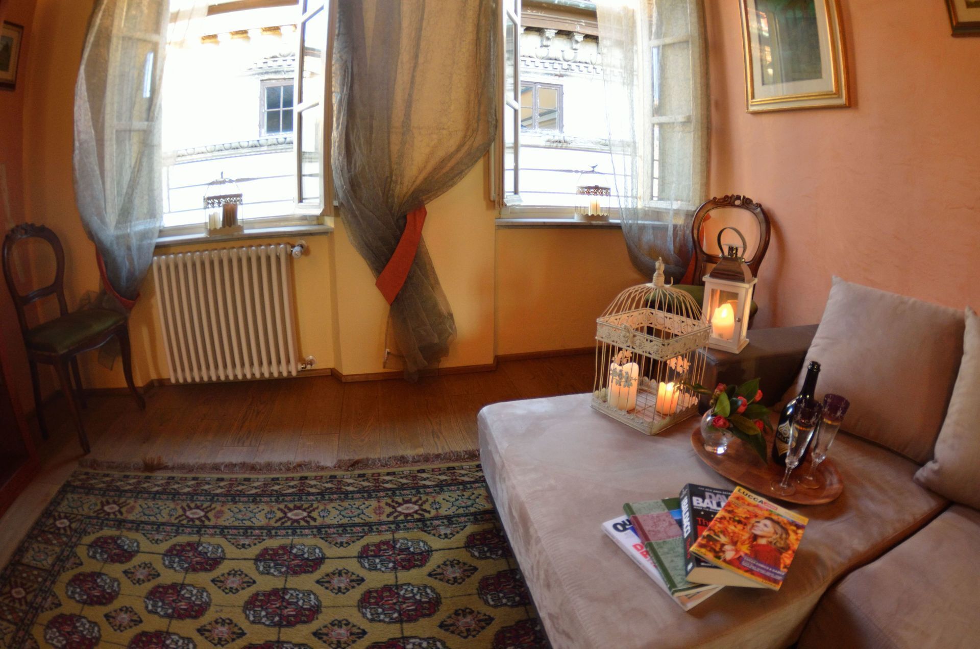 A cozy living room with a sofa, a small table with books and a candle, a window with curtains, and a radiator.