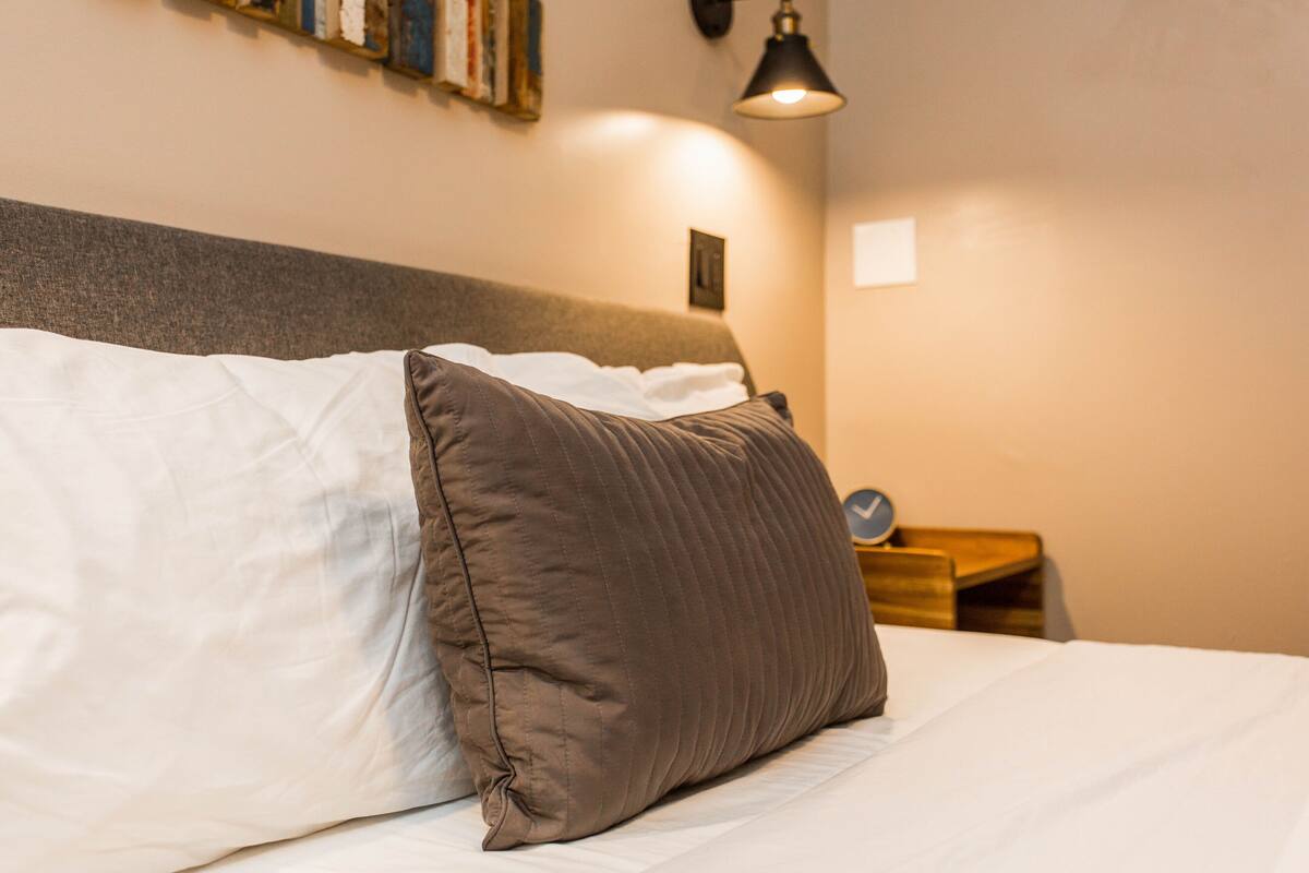 A neatly made bed with a white and brown pillow, a small bedside table with a clock, and a wall-mounted lamp.