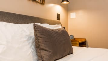 Un lit bien fait, avec un oreiller blanc et marron, une petite table de chevet sur laquelle se trouve une horloge, et une lampe fixée au mur.