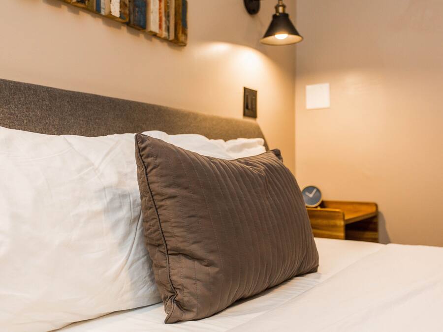 A neatly made bed with a white and brown pillow, a small bedside table with a clock, and a wall-mounted lamp.