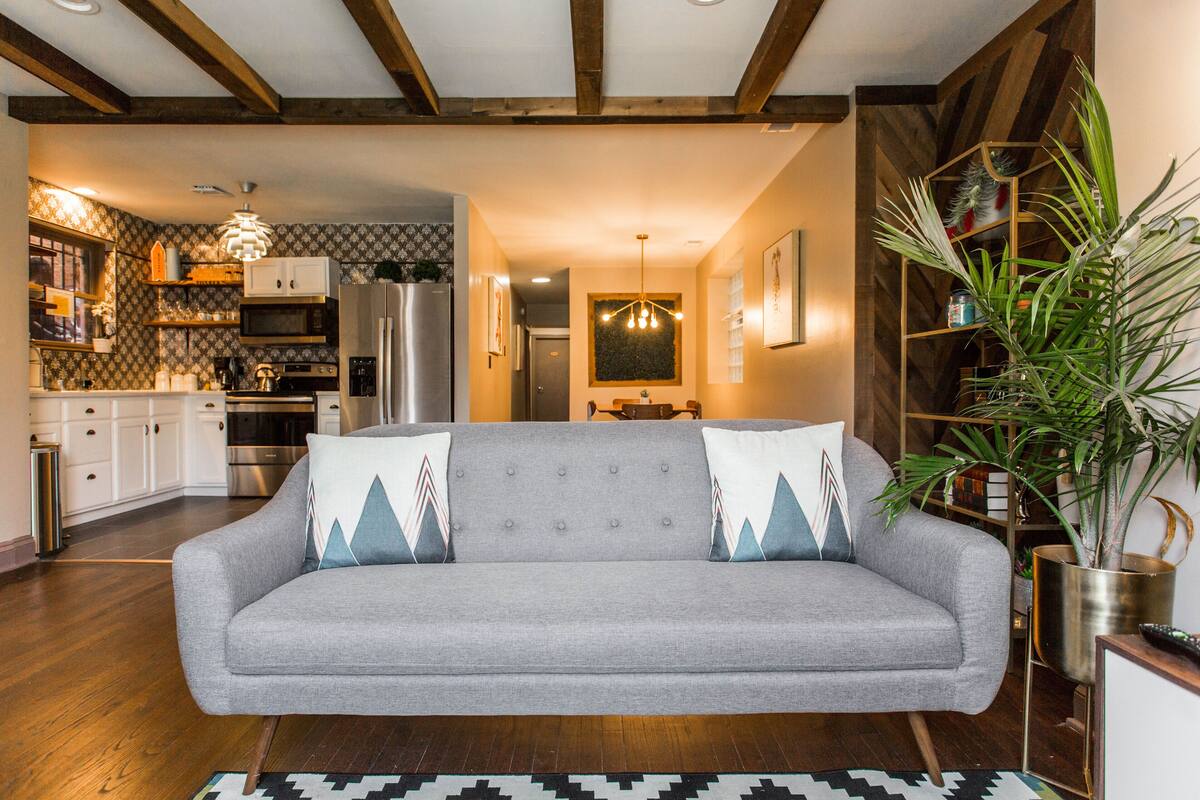 A living room with a grey sofa, wooden beams, a kitchen in the background, and a large plant.
